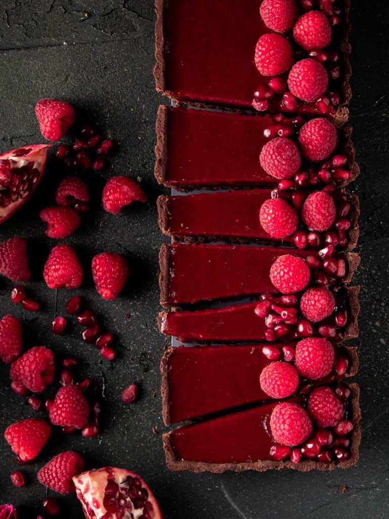 Chocolate Pomegranate Tart close up on dark board with raspberries on top and on the board.