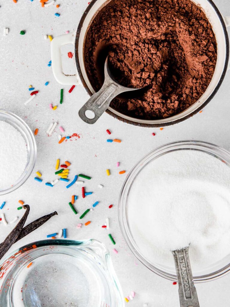 Bowls filled with cocoa powder, sugar and salt with a cup of water.