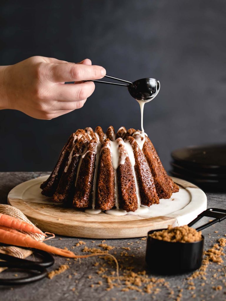 Glazing Carrot Bundt Cake on plate with measuring cup filled with sugar in front.