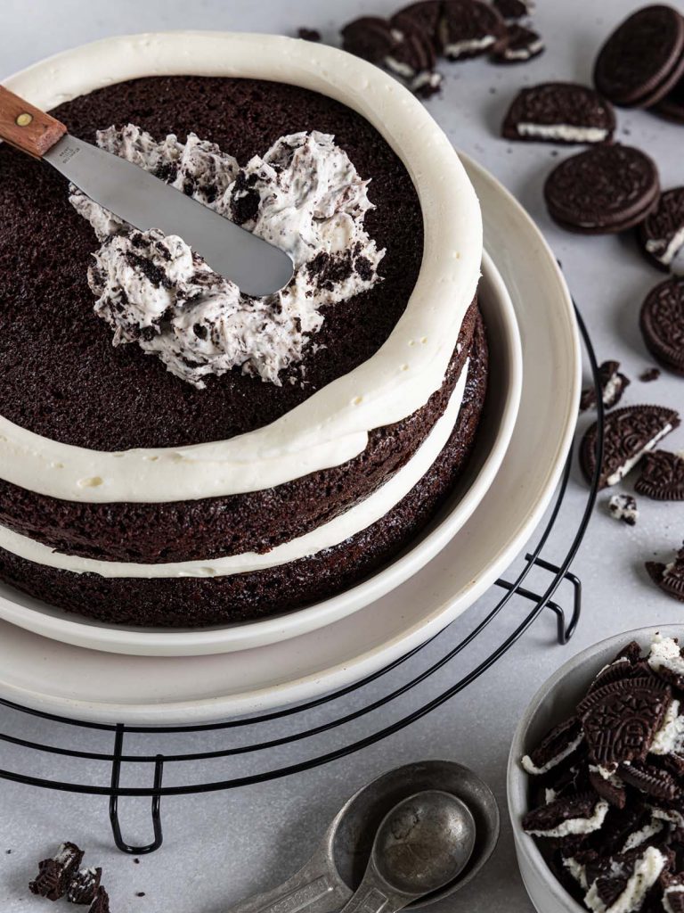 Stacking layers of chocolate cake with spreading filling on top with crushed Oreos around.