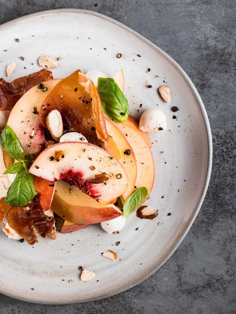 Salad on plate up close with sliced peaches, mozzarella, and fresh basil leaves.