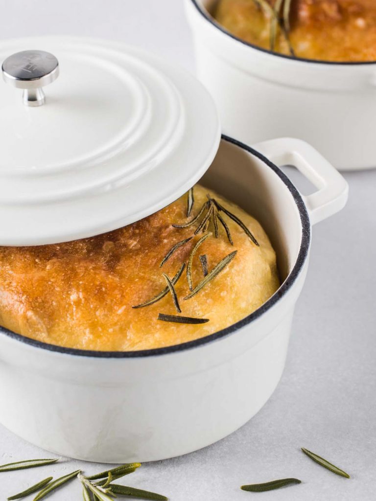 Focaccia bread at an angle in pan with lid and leaves of rosemary.