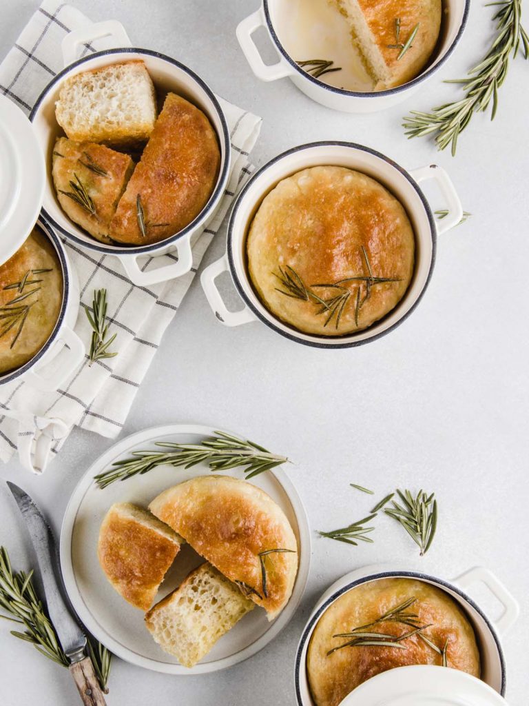 6 Mini focaccia breads in pans and on dish with sprigs of rosemary.