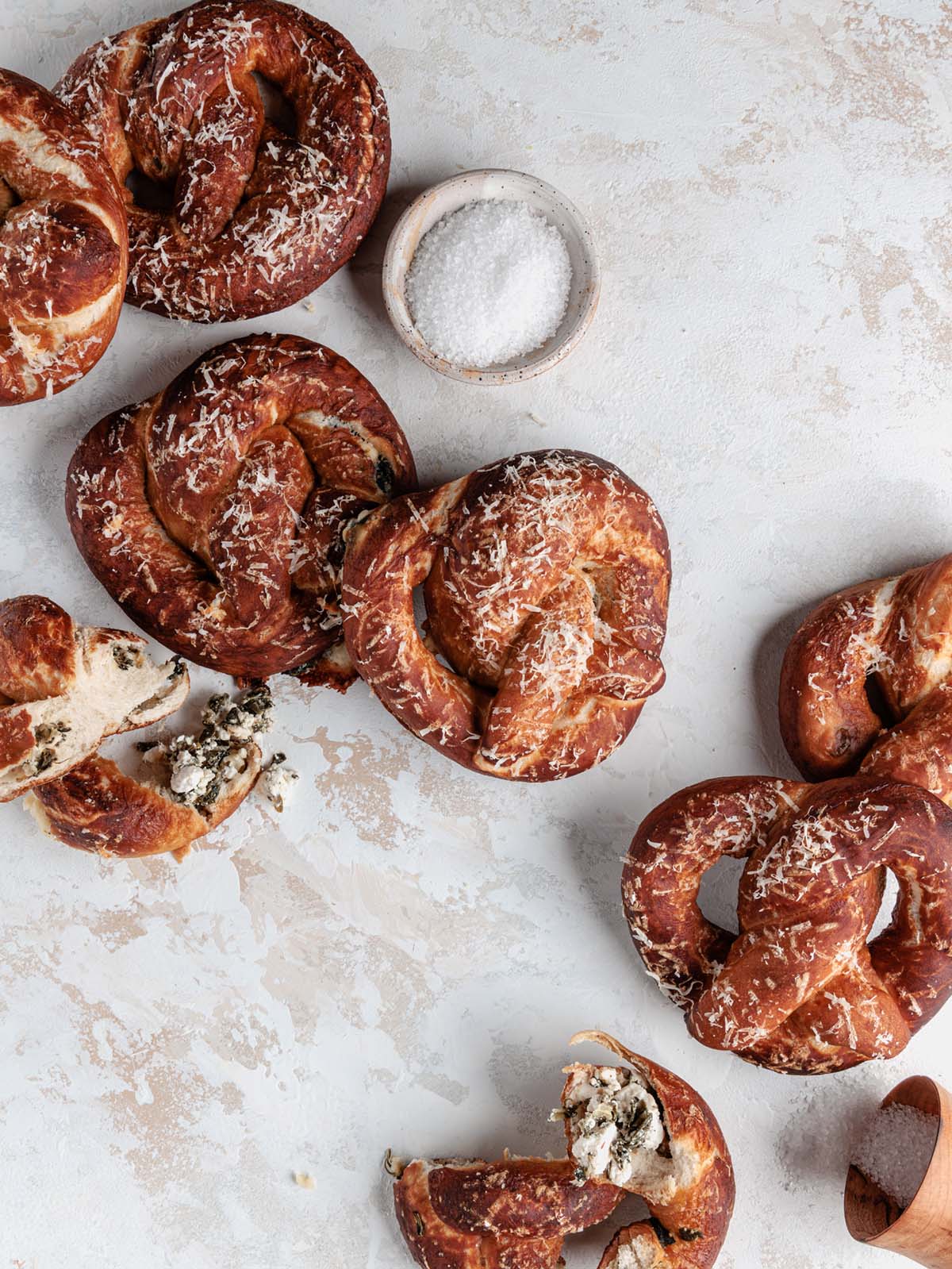 Soft pretzels scattered on board with some torn and container of salt spilling.