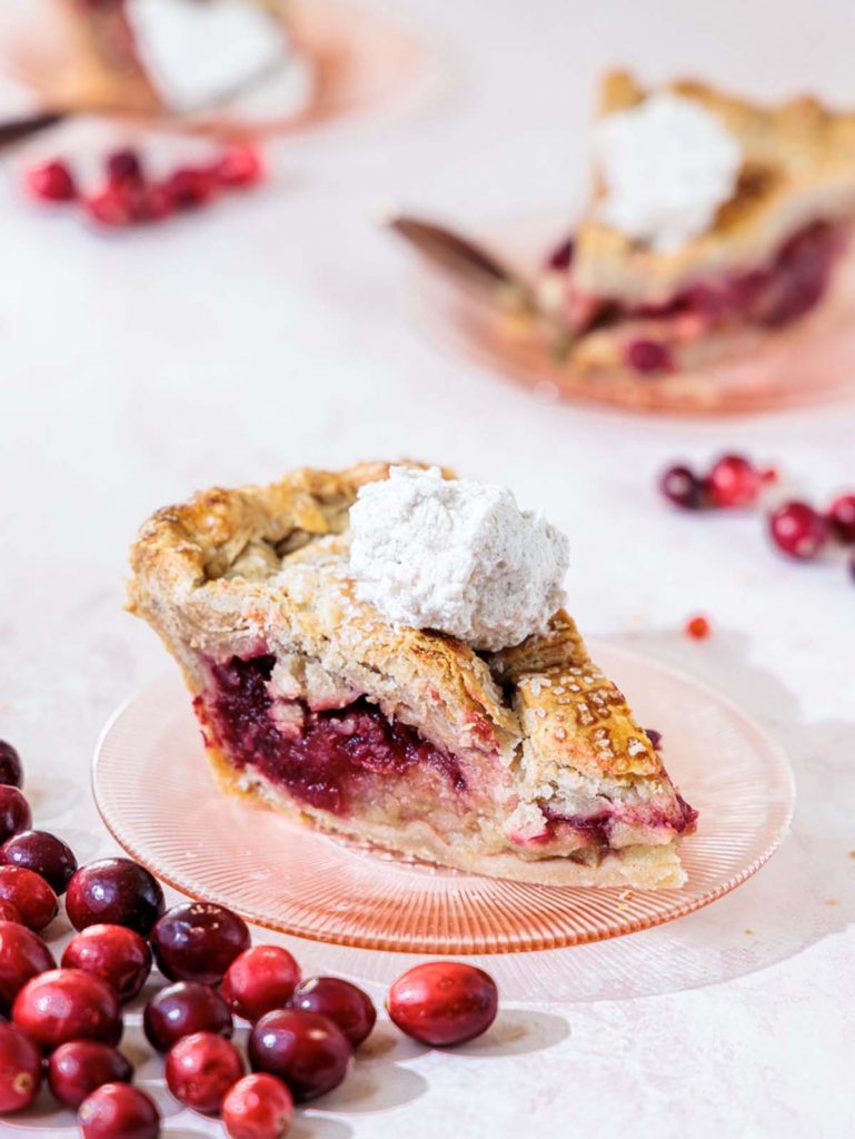 Side view of a slice of pie with scoop of whipped cream on top and cranberries scattered around.
