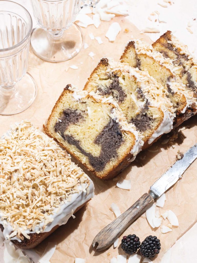 Sliced loaf cake on table with glasses and knife with toasted coconut on top.