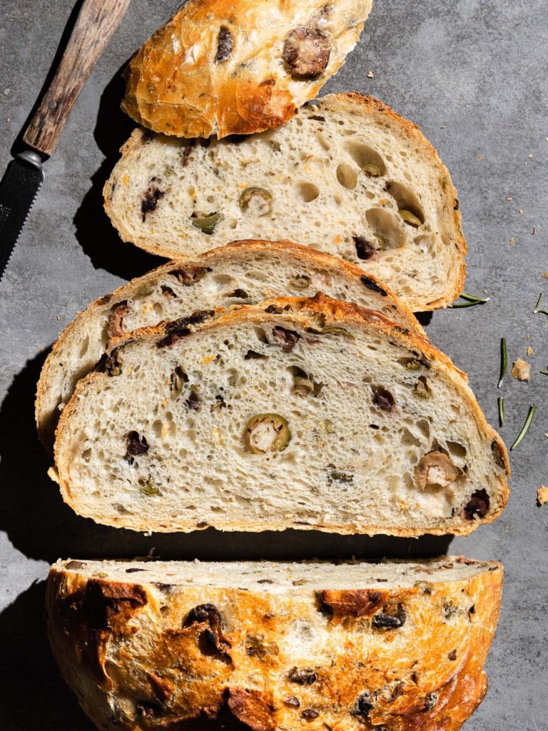 Half loaf of olive bread sliced on board with rosemary scattered.