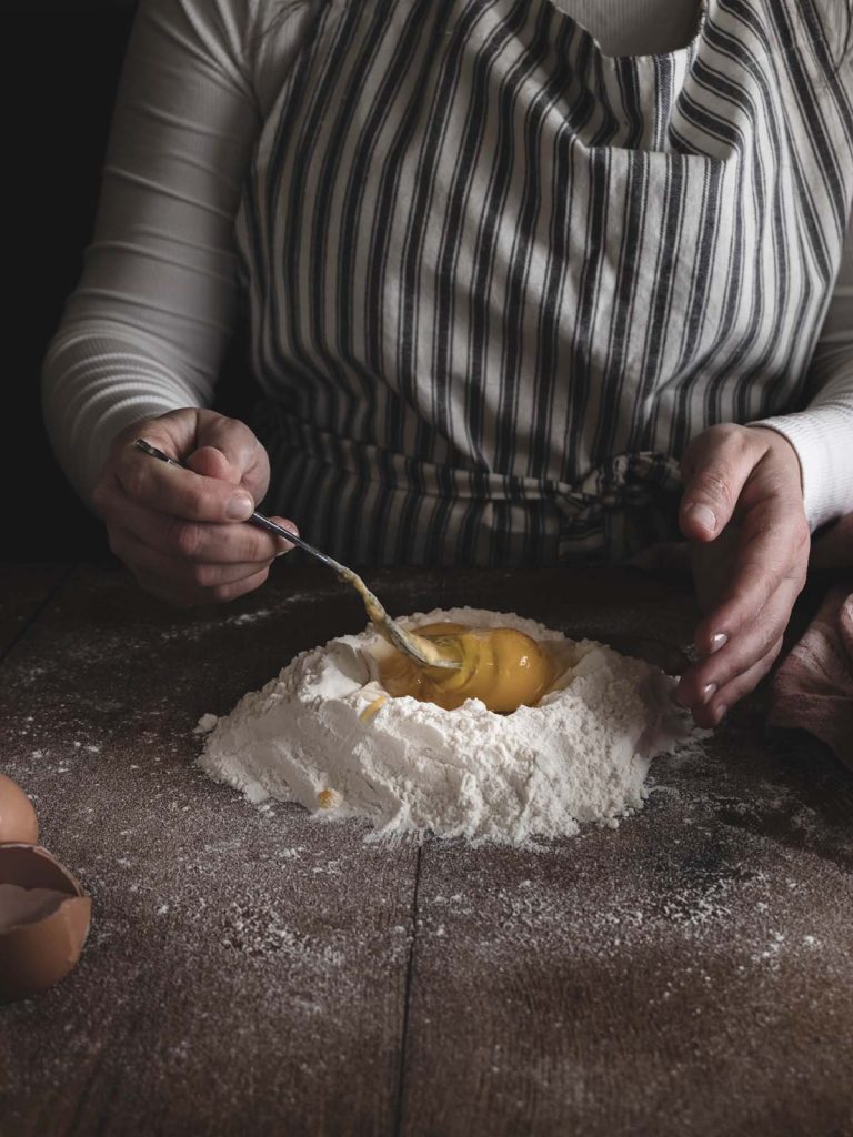 Chef beating eggs with fork in flour on counter.