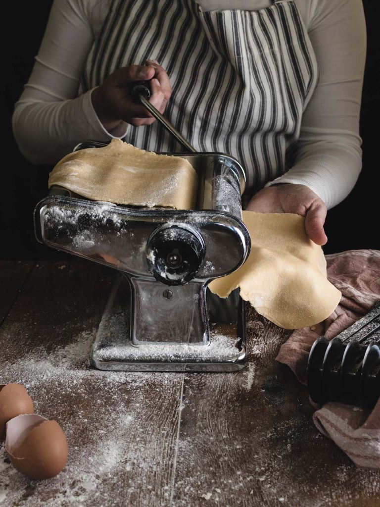 Chef rolling dough in pasta machine with egg shells and flour covered counter.