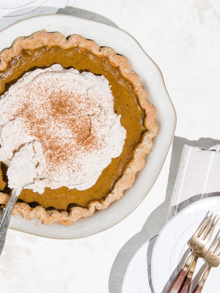 Pumpkin Pie in dish with forks and plate and spoon spreading whipped cream.