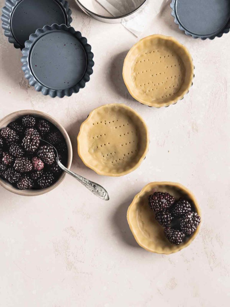 Tarts pans being lined with dough and bowl of blackberries.