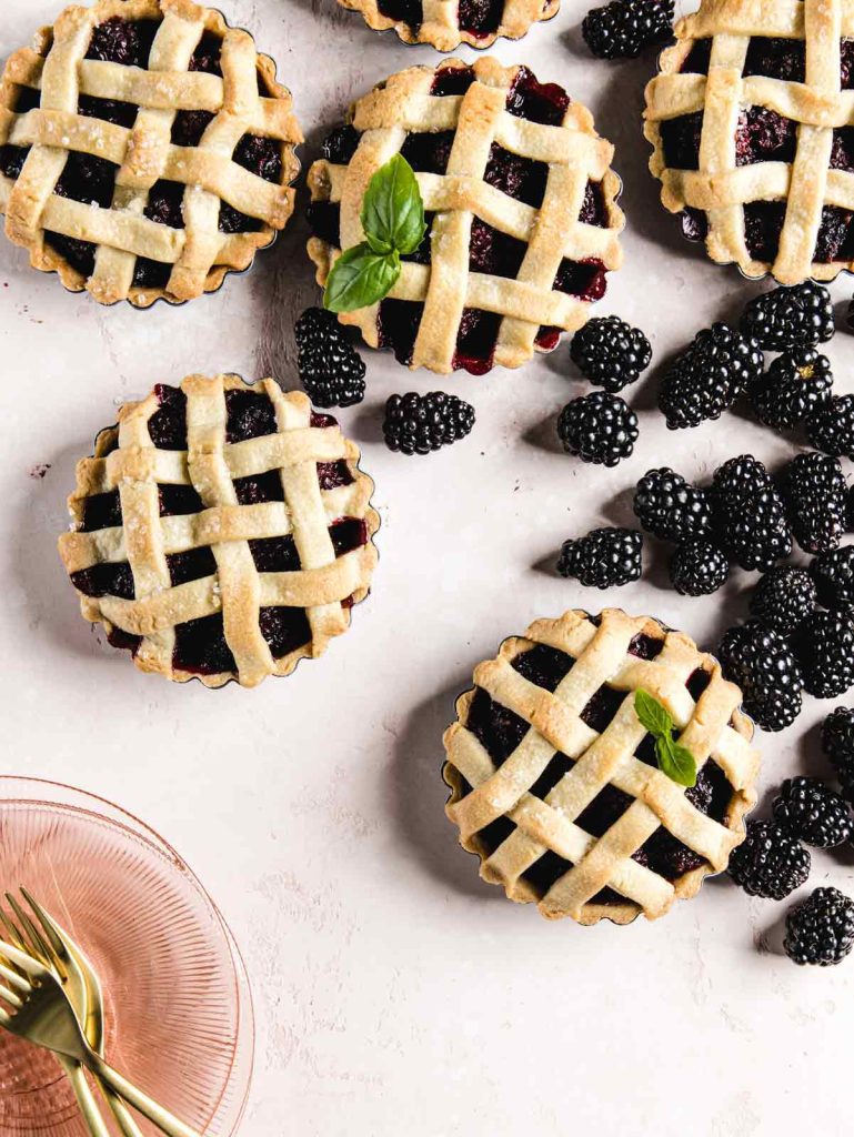 6 lattice topped tarts on pink board with scattered blackberries and pink plates