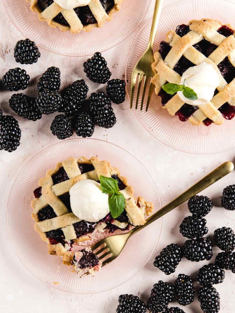 3 tarts on pink plates with ice cream on top and blackberries scattered on table