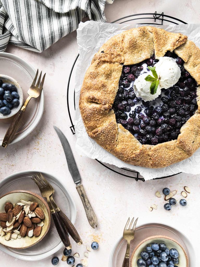 Galette on cooling rack surrounded by plates, silverware, blueberries and sliced almonds.