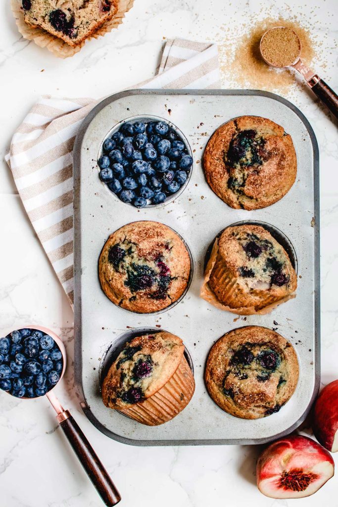 Blueberry Peach Muffins in muffin tin with fresh blueberries and bite removed from one muffin