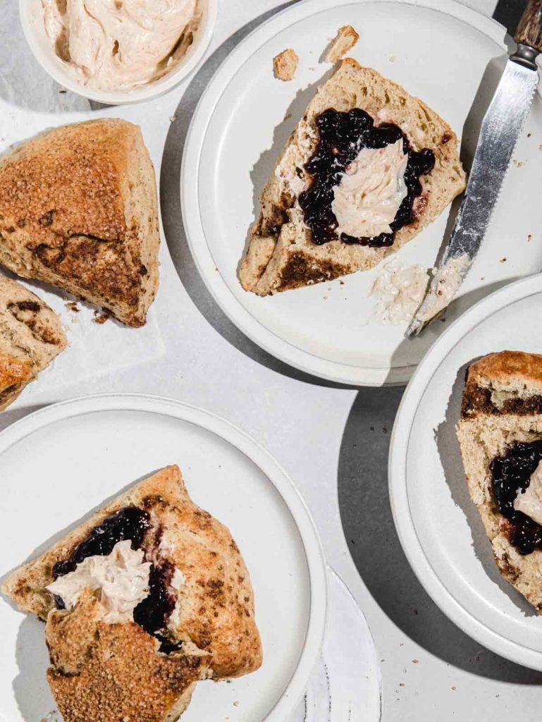 3 scones sliced with jam and butter with knife and whole scones on side.