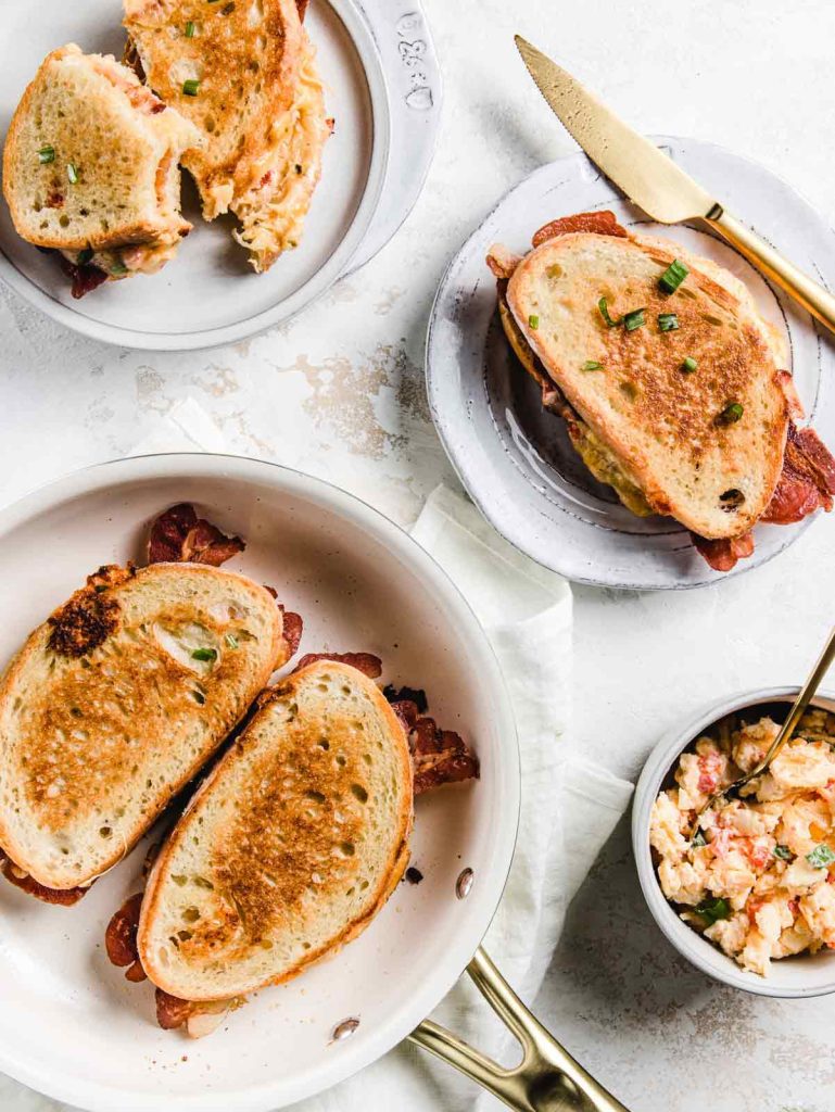 Multiple grilled cheese sandwiches on plates and in white skillet with bowl of pimento cheese.