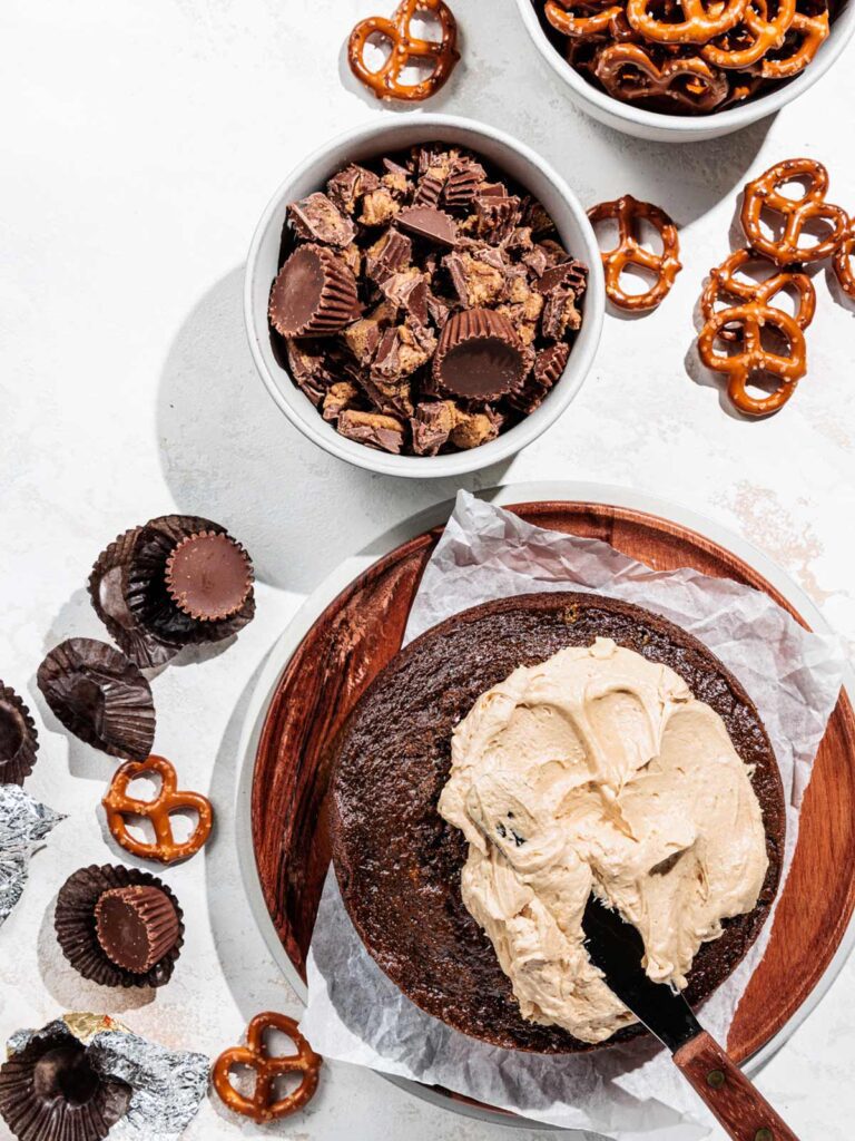 Chocolate cake being topped with peanut butter frosting with preztels and Reese's cups on table.