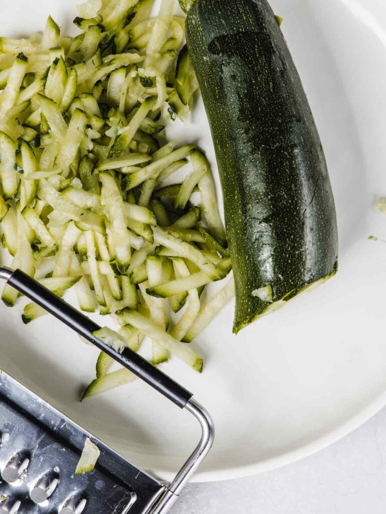 Grated zucchini on plate with grater and half zucchini.