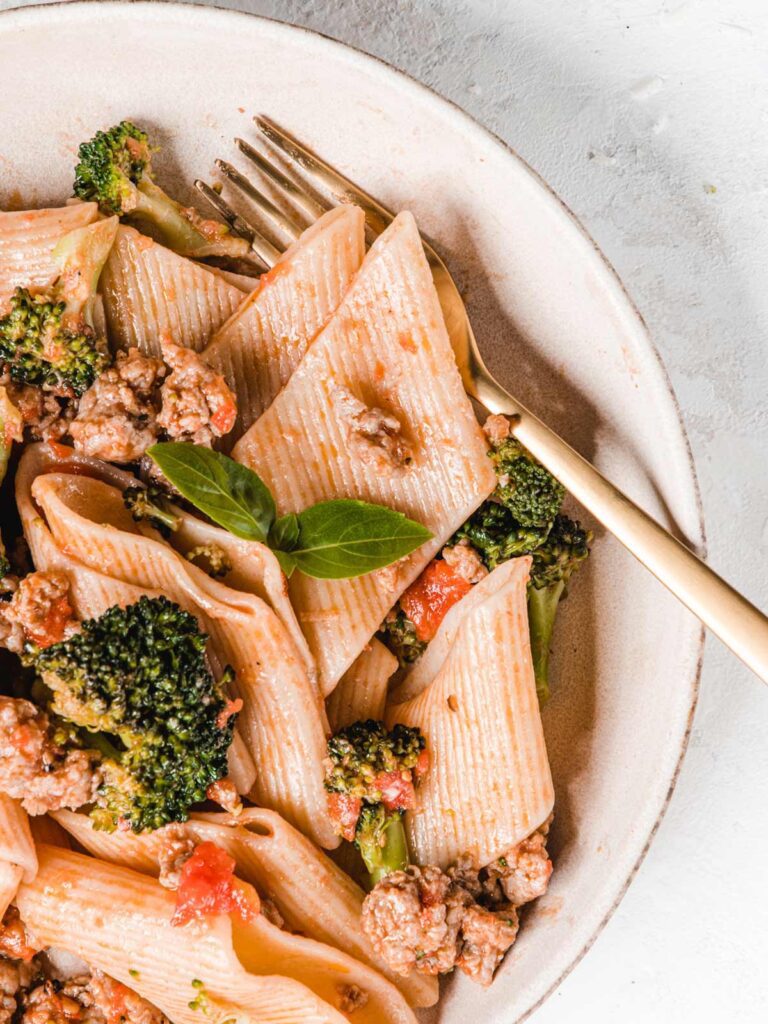 Close up of rigatoni on plate with broccoli and basil leave with fork on the side.