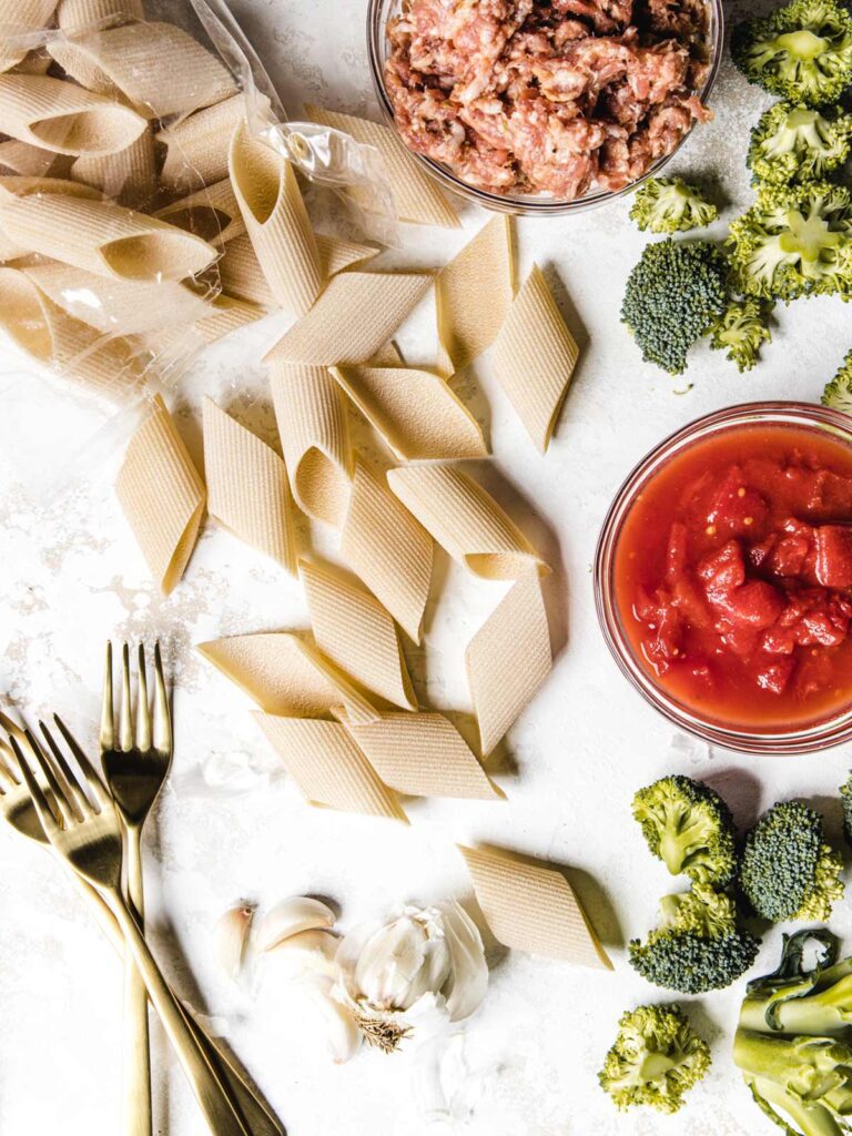 Pasta ingredients with dried pasta. tomatoes, broccoli and sausage on board.