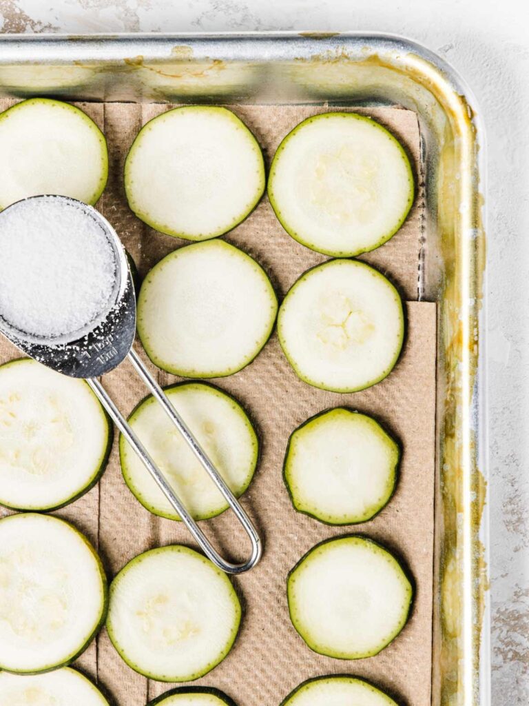 Sliced zucchini on baking sheet and paper towels with spoon of salt.