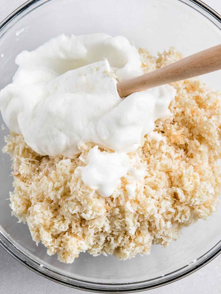 Bowl of coconut mixture with egg whites and spatula.