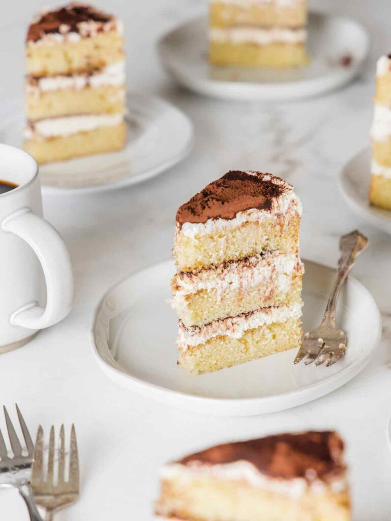 Multiple standing slices of tiramisu cake on plates with fork and coffee mug.