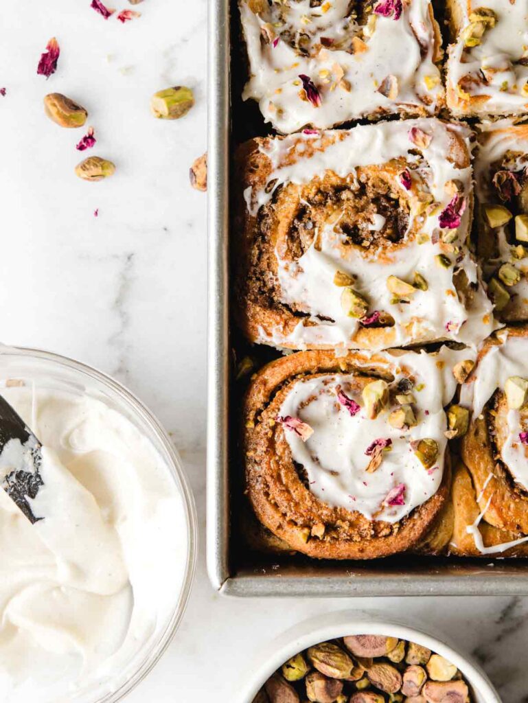 Close up of baking sheet with pistachio rolls and bowl of icing and pistachios.