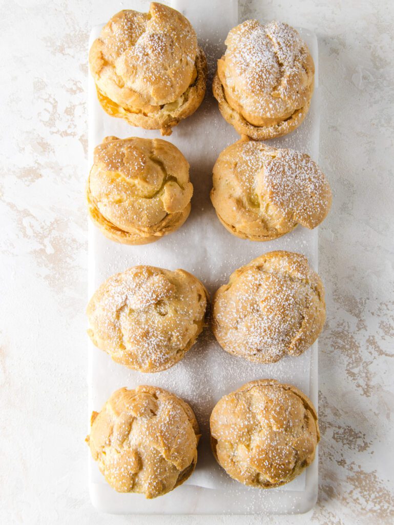 8 cream puffs on marble board with powered sugar on top.