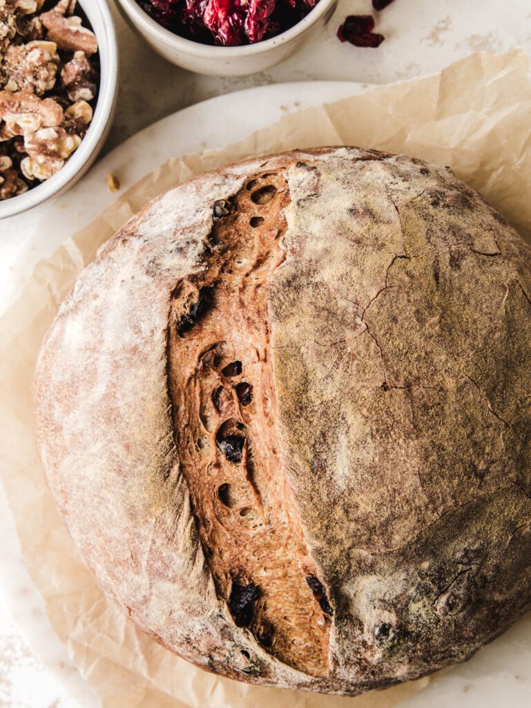 Crusty bread on parchment with bowl of walnuts and cranberries.