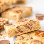 Stack of blondies on table with peanut butter cups and pretzels.