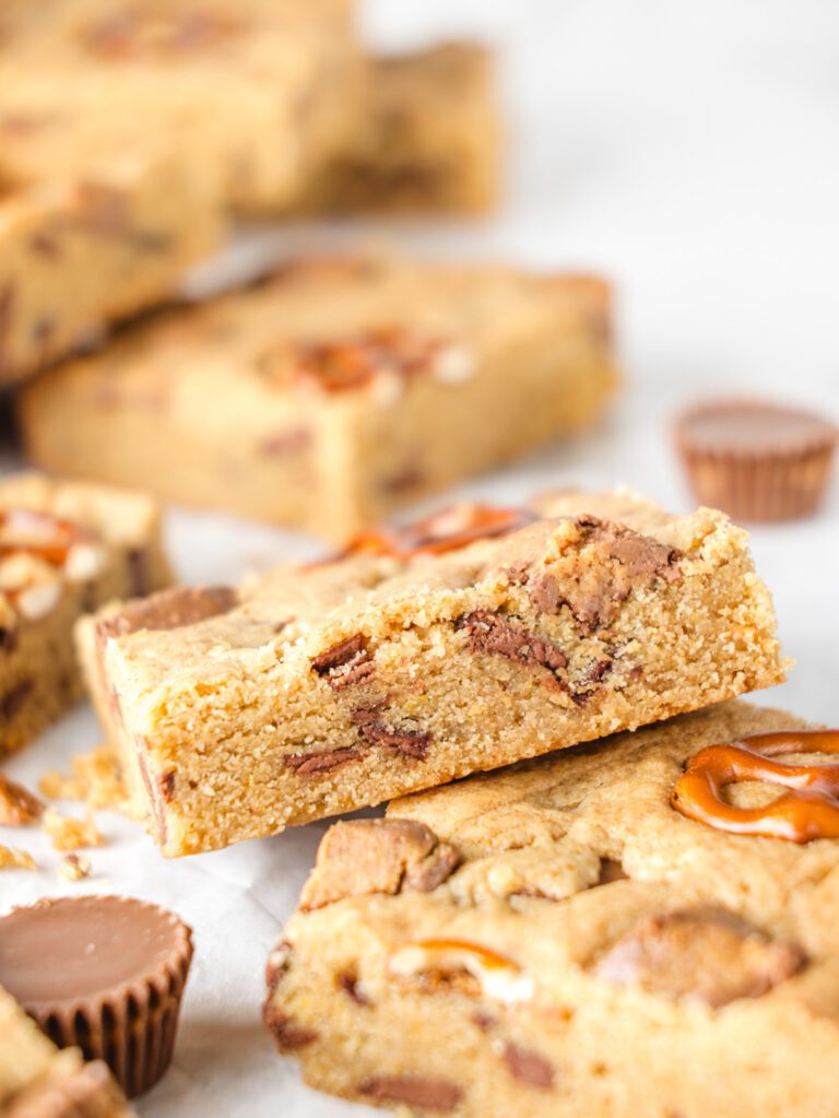 Stack of blondies on table with peanut butter cups and pretzels.